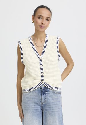 Femme portant un gilet en tricot crème sans manches avec des rayures bleu marine et des boutons, associé à un jean taille haute bleu clair sur un fond uni.