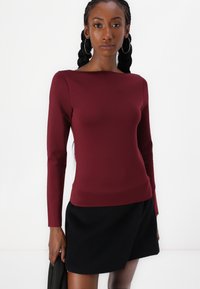 Haut à manches longues bordeaux en tissu lisse et extensible ; design ajusté avec un décolleté bateau associé à une jupe noire.