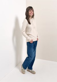 Lichtbeige textuur hoodie met een trekkoord, gecombineerd met blauwe wijde jeans. Schoenen zijn grijze sportschoenen met witte accenten.