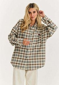 Blondynka w dużej, brązowo-szarej koszuli w kratę z guzikami i jasnych spodniach, pozująca z jedną ręką dotykającą włosów.