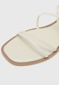 Sandale albe din piele ecologică cu trei curele subțiri, vârf pătrat și o talpă din cauciuc texturat de culoare maro. Design minimalist și plat.
