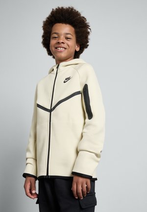 Hellbeige Nike Hoodie mit Reißverschluss, schwarzen Akzenten und einer Reißverschlusstasche am Ärmel. Bietet ein minimalistisches Design und eine weiche Textur.