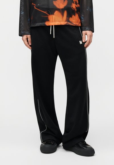 Personne portant un pantalon noir à jambes larges avec un liseré blanc sur les côtés, des baskets noires et un haut en maille noir et orange.