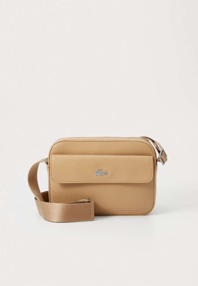 Beige rektangulær crossbody-veske med strukturert overflate, lomme med klaff på fronten, sølvfarget logoemblem og justerbar satengstropp på hvit bakgrunn.