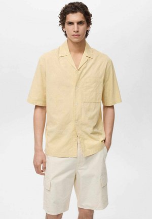 Kortærmet, knap-ned skjorte i blødt gult med et struktureret mønster. Båret med lyse beige cargo-shorts, der har sidelommer.