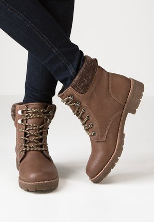 Botas de cuero marrón con cordones y puños de punto, usadas con vaqueros ajustados azul oscuro, un pie apoyado completamente y el otro ligeramente levantado.