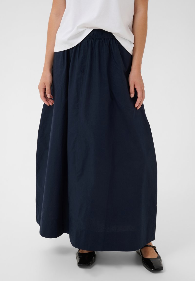 Femme portant une longue jupe bleu marine, un haut blanc et des chaussures plates noires, debout devant un fond uni.