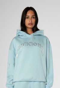 Sudadera azul claro hecha de una mezcla de algodón, con texto plateado brillante y adornos en las mangas. Corte holgado con un bolsillo tipo canguro.
