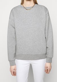 Sudadera gris con cuello redondo, puños y dobladillo acanalados. Hecha de tela suave, con una superficie ligeramente texturizada. Pantalones blancos visibles.