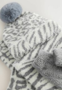 Grå och vit strukturerad mössa med en blå fuskpäls-pompon, som visar ett zigzag-mönster. Mjukt, fluffigt tyg som ger värme.