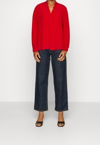 Rote Bluse mit langen Ärmeln und Schalkragen, kombiniert mit dunkelblauen weiten Jeans und schwarzen offenen High Heels. Glattes Material mit minimalen Details.