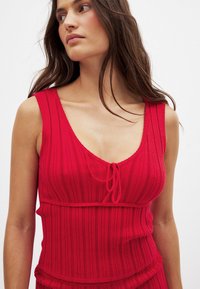 Red ribbed tank top featuring a deep V-neck and adjustable tie detail at the front. The fabric shows a subtle texture with vertical stripes.