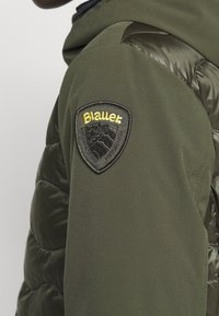 Olijfgroene jas met een mix van matte en glanzende texturen. Heeft een zwart geborduurd patch met "Blauer" en een bergontwerp op de schouder.