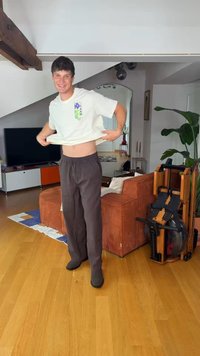 Jeune homme soulevant un t-shirt blanc, souriant, debout dans un salon avec un sol en bois, un canapé orange, une plante d’intérieur, un rameur et une télévision.