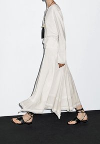 Kevyt, laskeutuva kermainen hame, jossa on laskokset; yhdistetty löysään kermanväriseen pitkähihnaiseen toppiin. Mustat remmikkäät tasapohjaiset sandaalit viimeistelevät asun.