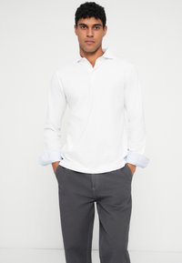 Balta ilgomis rankovėmis polo marškinėliai su apykakle ir šviesiai mėlynais rankovių akcentais, derinami su pilkais kelnais. Medžiaga atrodo glotni ir su laisvu siluetu.