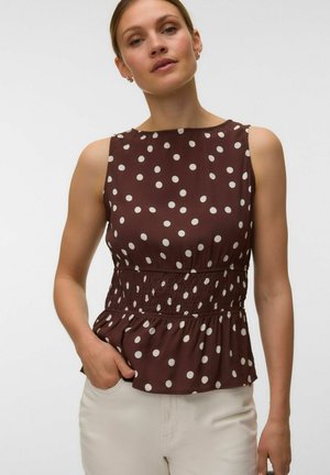 Femme portant un haut marron sans manches à pois blancs, taille smockée, associé à un pantalon clair, debout devant un fond uni.