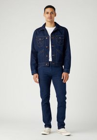 Chaqueta de mezcla de algodón azul oscuro con cierres de botón, dos bolsillos en el pecho y un diseño estructurado, combinada con jeans oscuros a juego y zapatillas claras.