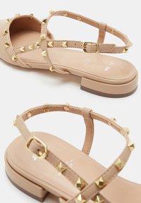 Sandalias beige con un tacón bajo, con tachuelas doradas a lo largo de las correas, cierre de hebilla y una textura superior de plástico suave.