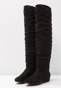Botas negras sobre la rodilla hechas de un material suave similar al ante, con una textura arrugada y punta redonda, y un tacón cuña bajo.