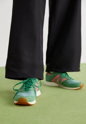 Grüne Wildleder- und Mesh-Sneaker mit rosa Akzenten, weißer Zwischensohle und braunem Gummiaußensohle, kombiniert mit schwarzen Hosen auf einem grünen Boden.