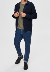 Marinblå ribbad cardigan, grön crew neck t-shirt under, blå taperade jeans och svarta snörskor med uppvikta byxben.