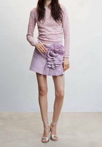 Roze kanten top met lange mouwen, gecombineerd met een lavendelkleurige geplooide minirok met een grote ruchesversiering, gedragen met zilveren sandalen met bandjes.