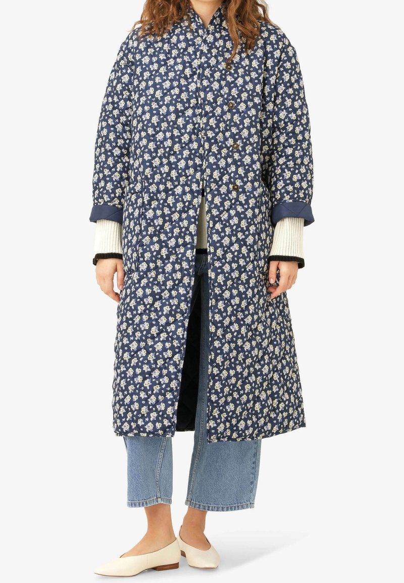 Femme portant un long manteau matelassé bleu marine à motifs floraux, un jean court bleu clair, des manches de pull crème visibles, et des chaussures blanches plates.