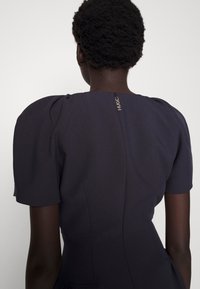 Dunkelgraues Kleid mit kurzen, puffigen Ärmeln, das über eine Nahtdetails und einen Reißverschluss auf der Rückseite mit "HUGG"-Branding an der Oberseite verfügt. Glatte Textur.