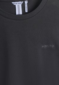 Crna majica opuštenog kroja napravljena od glatkog materijala, s malim reljefnim logotipom "MORATO" na lijevoj strani grudi i okruglim dekolteom.