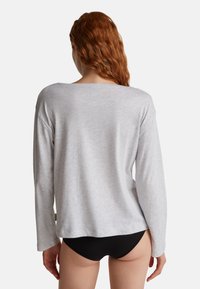 Frau mit langen roten Haaren, die ein hellgraues langärmliges Oberteil und schwarze Unterwäsche trägt, steht mit dem Rücken zu einem weißen Hintergrund.