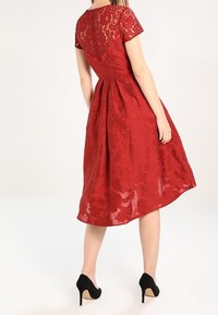 Robe en dentelle rouge avec des manches courtes, buste ajusté, jupe évasée, motif floral et un dos nu. Portée avec des chaussures à talons hauts noires.