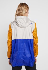 Färgglad vindjacka med grå, orange och blå paneler; har en huva och North Face-logotypen på övre delen av ryggen.