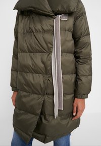 Olivgrön vadderad jacka med bred krage, med diagonalt framstängning och ett mönstrat slipsdetalj. Texturerad quiltad design.