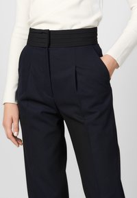 En person iklädd en åtsittande, ribbad vit topp och marinblå högmidjade byxor med en strukturerad midja, händerna i fickorna, visad från midjan och ner.