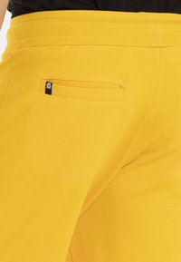 Gele sportieve shorts met een gladde textuur, voorzien van een achterzak en een klein zwart logo-accent. Elastische tailleband voor comfort.