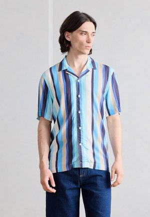 Άνδρας με σκούρα μαλλιά μέχρι τους ώμους, που φοράει κοντομάνικο πουκάμισο με κατακόρυφες μπλε, λευκές και μπεζ ρίγες, και σκούρα μπλε τζιν.