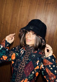 Pălărie neagră tip bucket cu margine cu imprimeu leopard și un emblemă, purtată împreună cu o cămașă colorată, cu model floral și geometric.