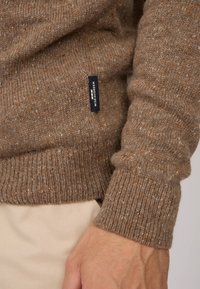 Pull en laine marron avec un ourlet et des poignets côtelés, parsemé de fibres plus claires et plus foncées. Comprend une étiquette de marque visible.