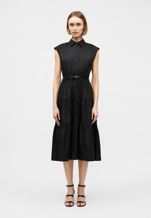 Γυναίκα στέκεται φορώντας μαύρο midi φόρεμα χωρίς μανίκια με κουμπιά, γιακά, ζώνη στη μέση και μαύρα ψηλοτάκουνα σανδάλια, σε λευκό φόντο.