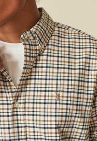 Een button-up shirt in een beige, bruine en marineblauwe ruitpatroon; heeft een geplooide kraag, een witte onderhemd en een klein geborduurd accent op de borst.