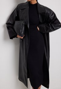 Manteau en cuir noir avec de grands revers surdimensionnés, associé à une robe en maille côtelée noire. Le mannequin tient une pochette noire tressée, avec une surface texturée.