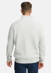 Mand iført en lysegrå ribbet turtleneck sweater og mørkeblå jeans, set fra ryggen mod en hvid baggrund.
