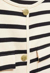 Gestreifter Cardigan in Creme und Schwarz, mit einem Knopfverschluss mit goldfarbener Akzentuierung und zwei vorderen Taschen, die mit weißem Garn genäht sind.
