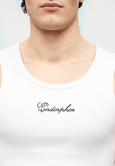 Gros plan sur une personne portant un débardeur blanc avec le mot « Endorphin » brodé en lettres noires cursives sur la poitrine.
