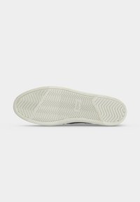 Semelle d'une chaussure noire avec une semelle extérieure en caoutchouc blanc montrant un motif chevrons et le nom de la marque "TOMS" sur fond blanc.