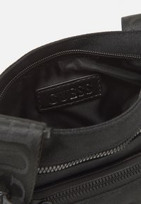 Intérieur d'un sac en nylon noir présentant une surface texturée, une étiquette logo GUESS et une poche zippée le long du bord.