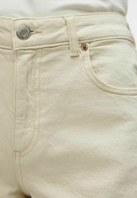 Jeans de mezclilla beige con un acabado texturizado, que presentan un cierre de botón de metal y detalles de costura sutiles en los bolsillos y la cinturilla.