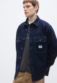 Σκούρο μπλε πουκάμισο από τζιν με κουμπιά στο μπροστινό μέρος, δύο τσέπες στο στήθος και διακριτή ανοιχτόχρωμη ραφή. Φορεμένο πάνω από ένα γκρι t-shirt.