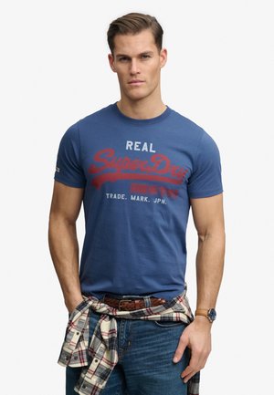 Mann trägt ein blaues SuperDry-T-Shirt, ein kariertes Hemd, das um die Taille gebunden ist, einen geflochtenen braunen Gürtel, blaue Jeans und eine Armbanduhr, steht mit den Händen in den Taschen.